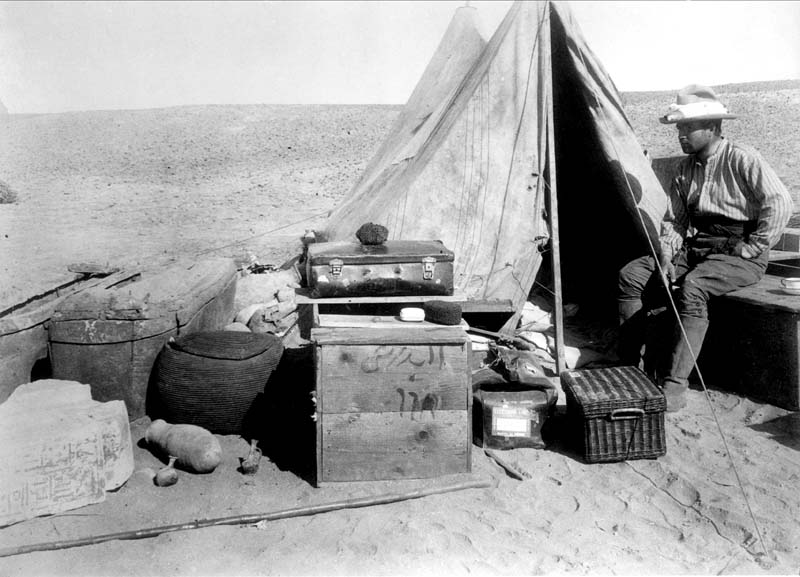 Black and white photograph of a man sitting beside a tent in the desert.
