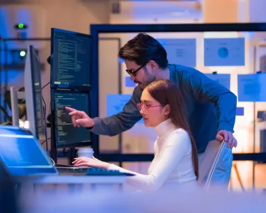 A man stands beside a seated woman working on a computer, both looking at a monitor with code in an office setting. A man stands beside a seated woman working on a computer, both looking at a monitor with code in an office setting.