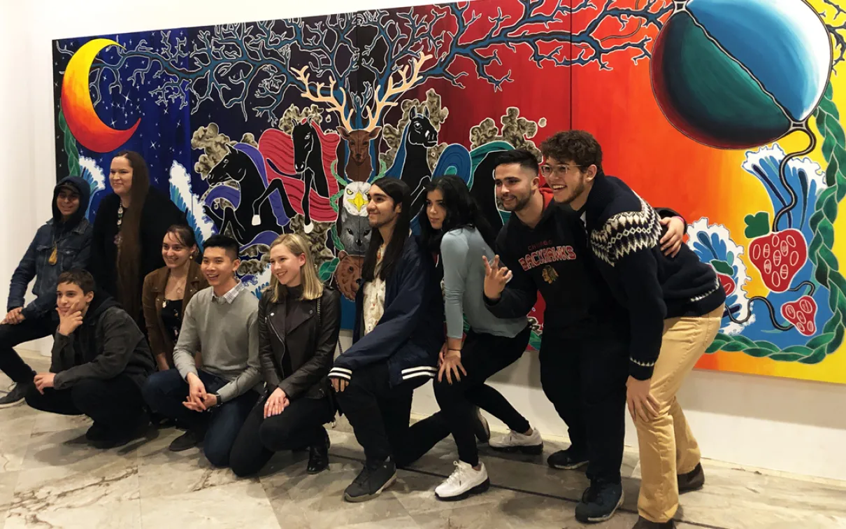 Nine Youth Cabinet members pose in front of a large mural on the wall with ROM Indigenous Knowledge Resource Teacher Leslie McCue.