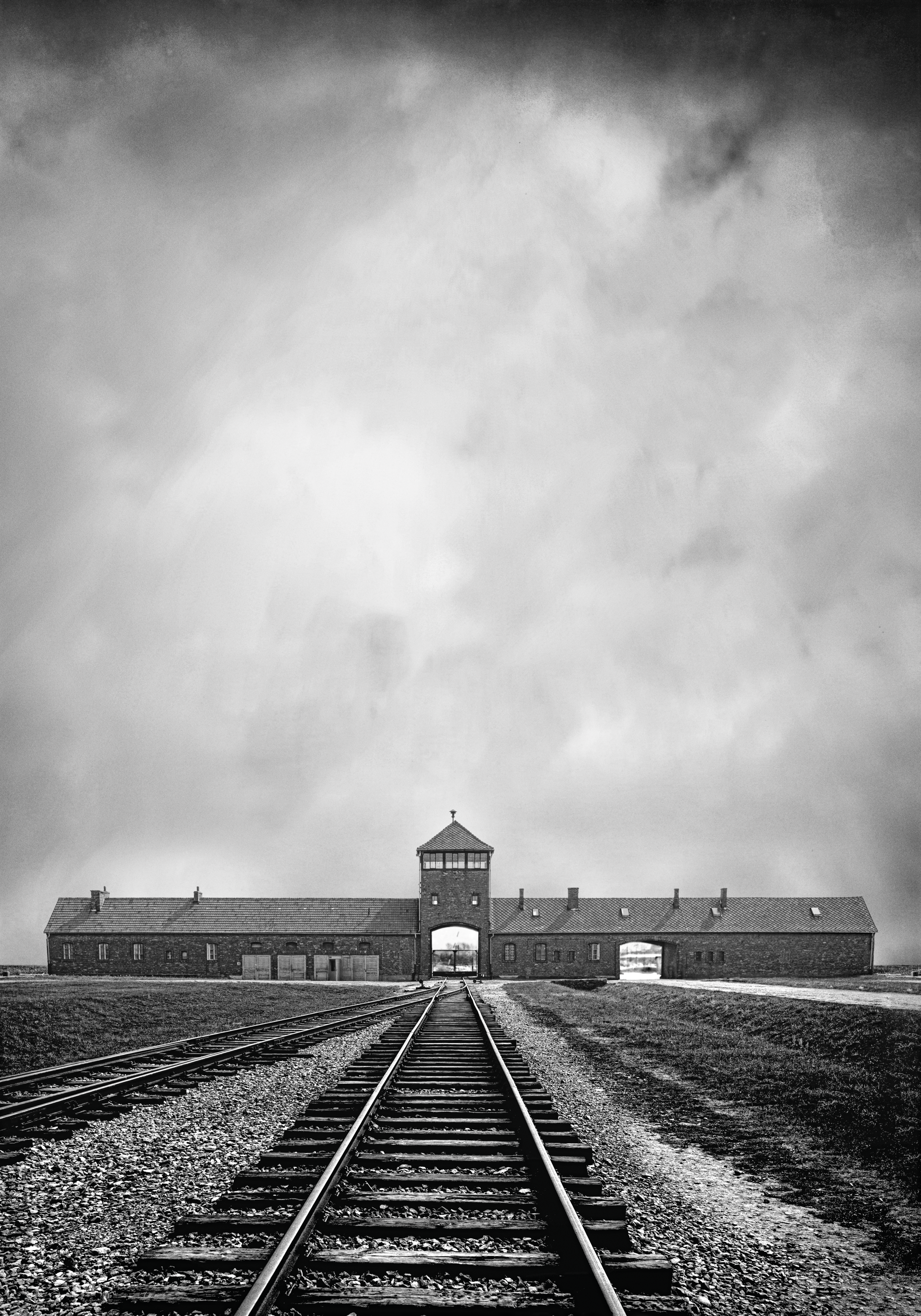 Black and white archival image showing train tracks in the foreground leading into the distance towards the entrance to the Auschwitz camp.