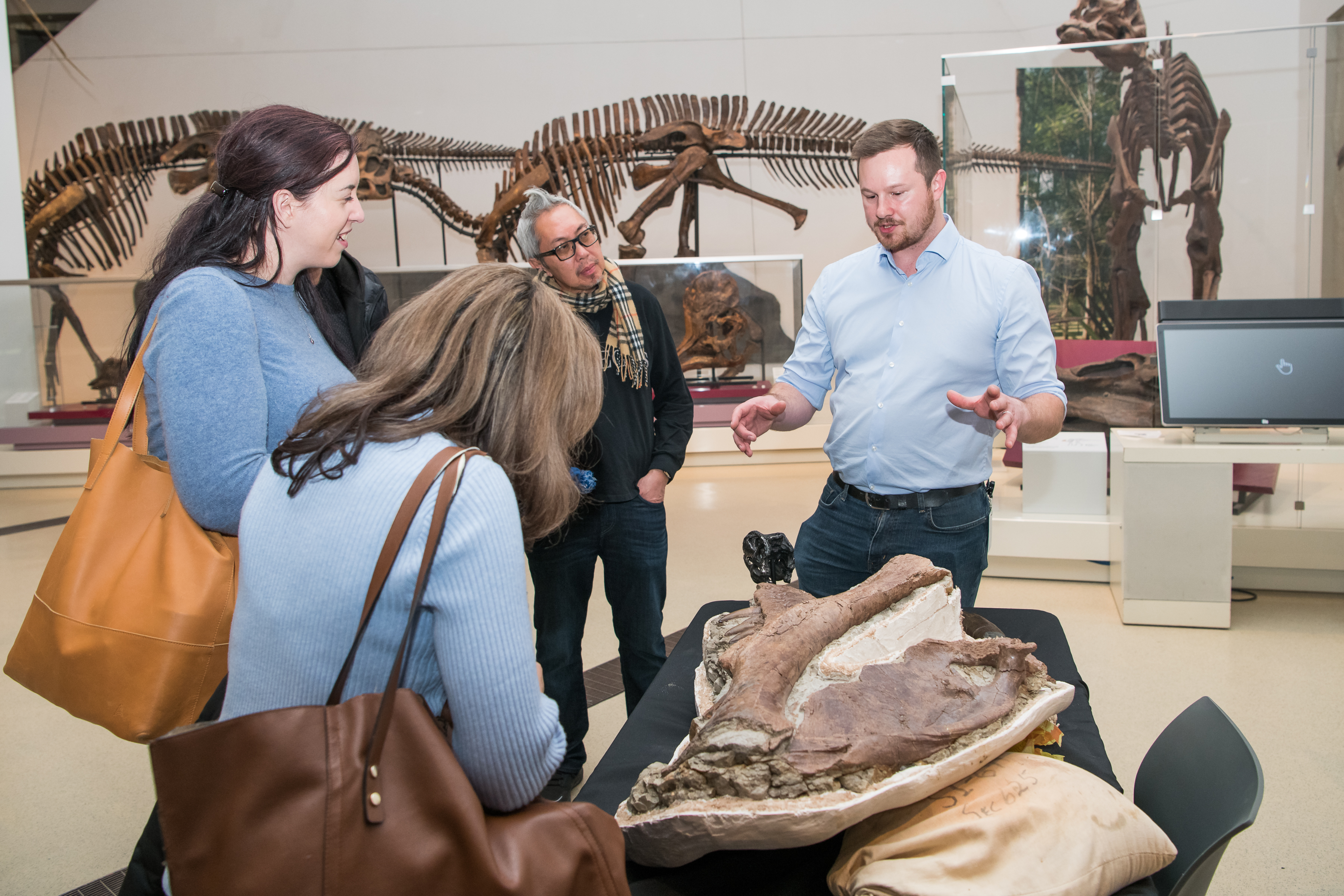 Three people look at a dinosaur bone while a ROM expert explains.