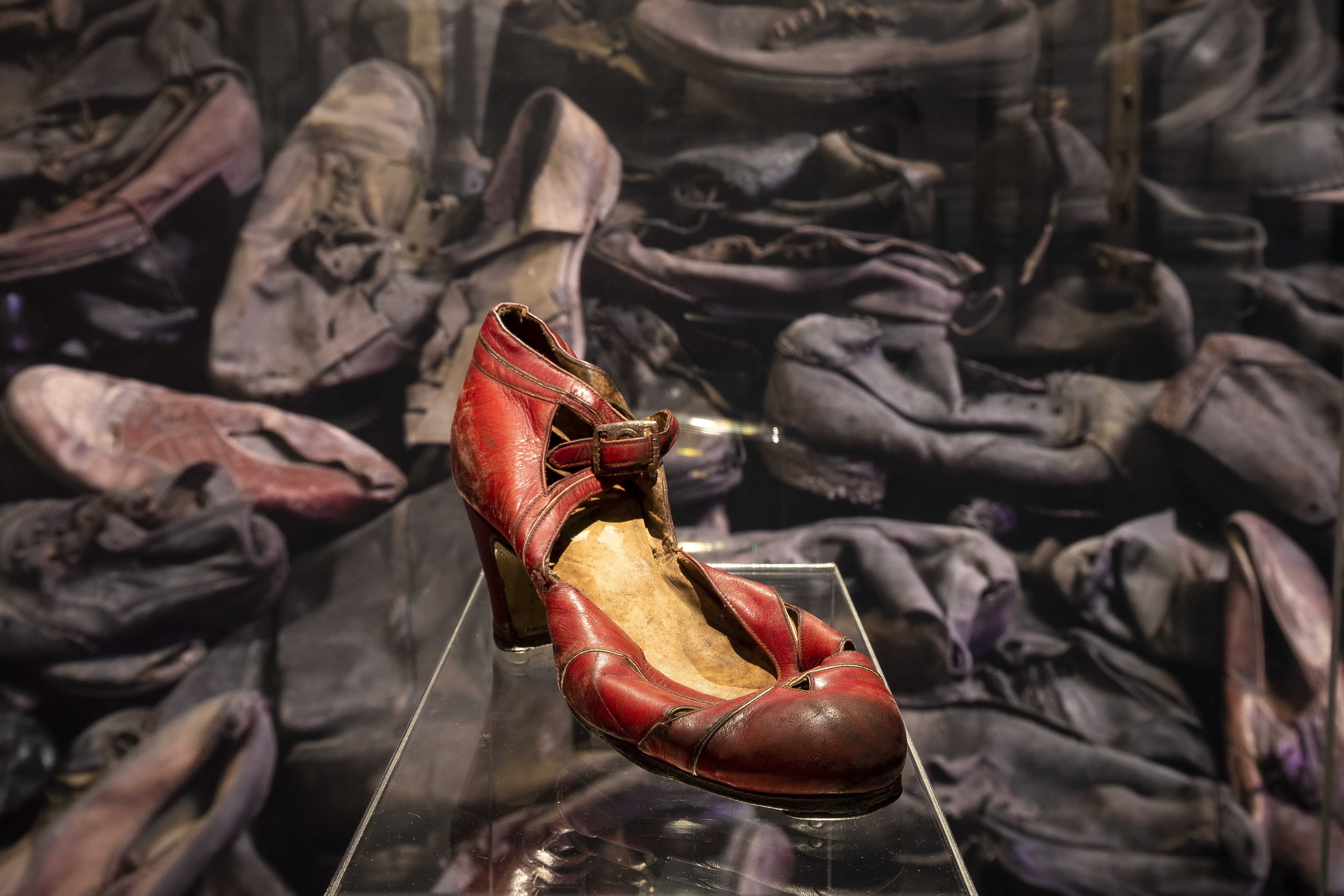 A battered and worn red high-heel shoe, shown in front of a pile of hundreds or other shoes.