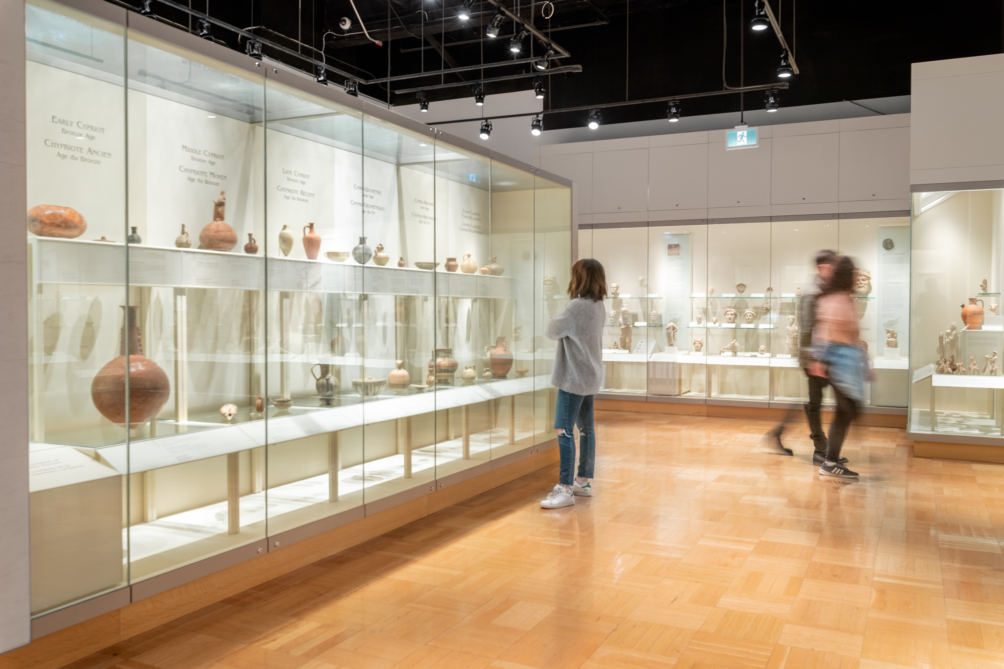 Three people walk around a gallery filled with ancient pottery in cases.