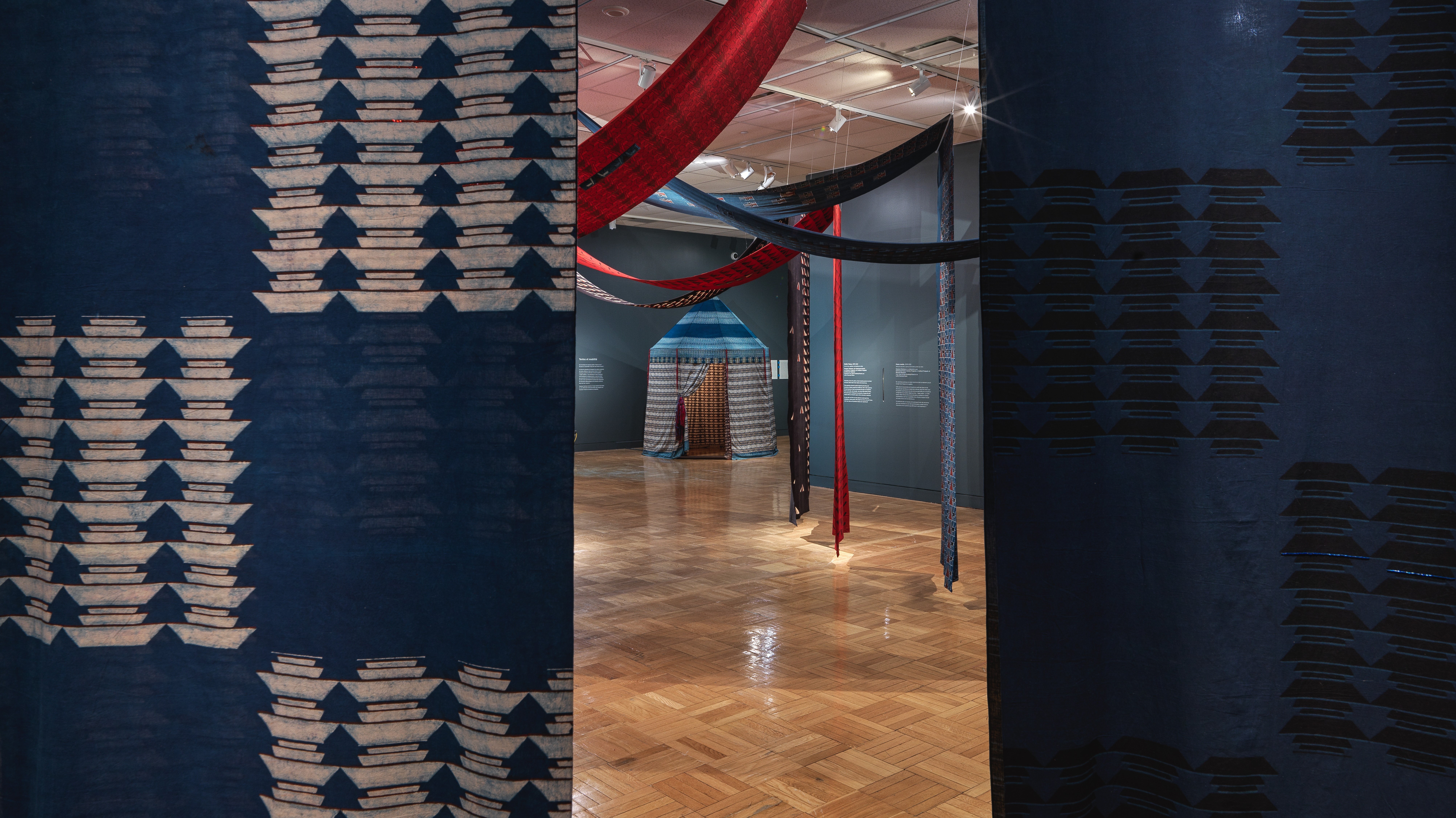 View of a cotton tent structure seen through a break in two printed, hanging cotton panels in the exhibition Swapnaa Tamhane: Mobile Palace, at ROM.