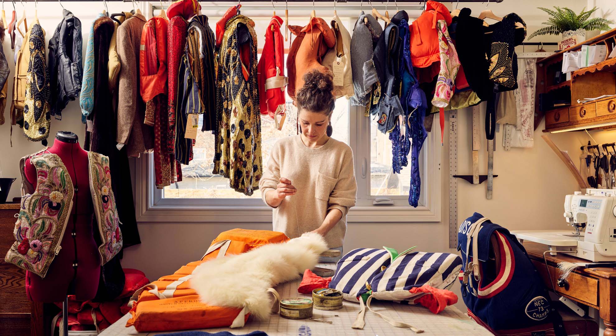 Image of the artist working at a large table containing works in progress, with a selection of lifejackets suspended along the wall behind.
