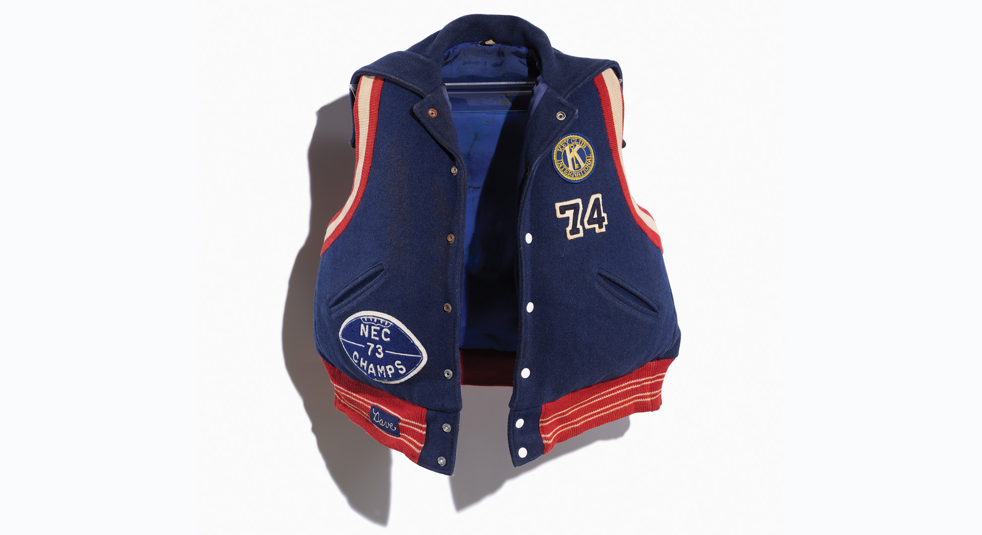 Image of a repurposed life jacket covered in a navy varsity jacket with orange and red trim and school letters and team patches/numbers. Image taken on a white background.
