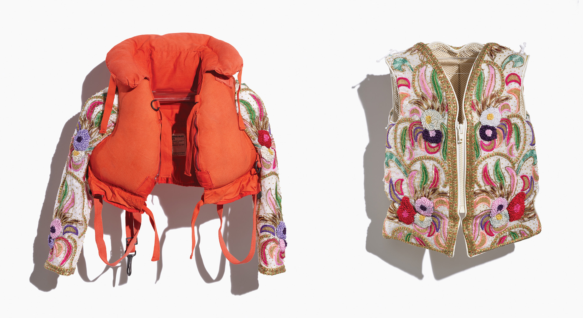 Image of an adult and a child's repurposed life jackets, the adult's completely covered, and the child's embellished with added sleeves, in a colourful beaded fabric depicting a floral pattern in reds, pinks, greens, and gold. Side-by-side image of both jackets taken on a white background.
