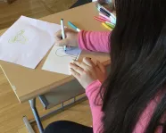 The back of a girl with long dark hair who is sitting at a desk and drawing an outline of a tree on a piece of paper with colourful markers.
