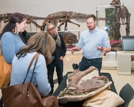 Trois personnes regardent un os de dinosaure pendant qu'un expert ROM explique.