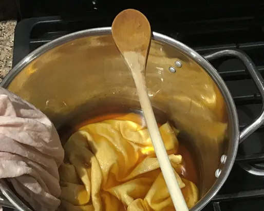 Pale pink shirt submerged halfway into amber dye in a stainless steel pot.
