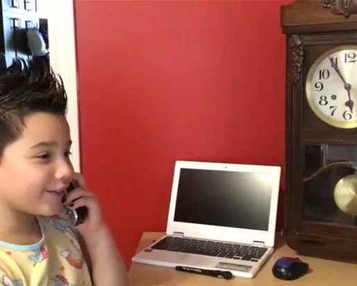 A girl with short spiky hair smiles while talking on the phone and sitting next to a laptop and grandfather clock on the table.
