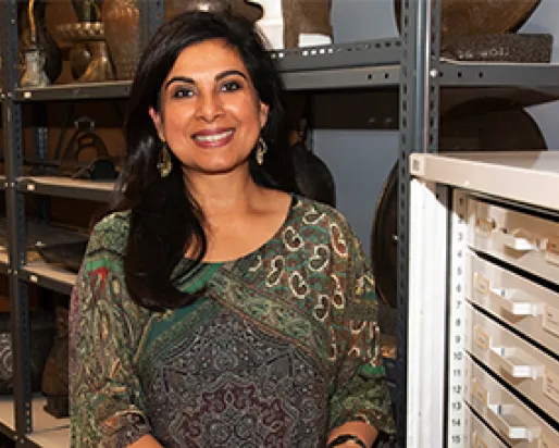 Curator Fahmida Suleman, a smiling woman with tan skin and long dark hair, wearing a colourful print shirt and dangly earrings, stands in front of metal shelving units holding metal Islamic urns and containers.
