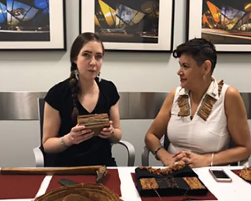 ROM staff Jessie Jakumeit and J’net Ayayqwayaksheelth hold a cedar block and sit at a table displaying four objects made using cedar bark.
