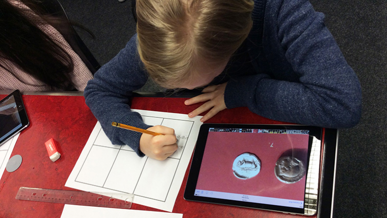 A student draws on a piece of paper with a coin image open on her iPad