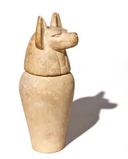 A sand-coloured Egyptian jar with a lid sculpted in the shape of a jackal's head.