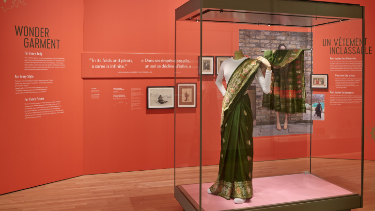 Installation présentant des panneaux de texte rouges derrière une vitrine contenant un sari de soie verte avec des fils métalliques tissés, porté sur un T-shirt blanc, sur un mannequin portant des chaussures de sport blanches.