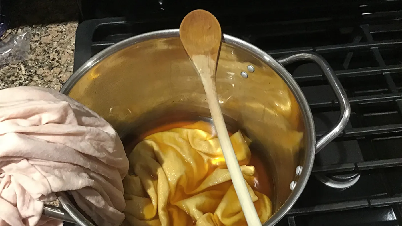 Chemise rose pâle immergée à moitié dans une teinture ambrée dans un pot en acier inoxydable.
