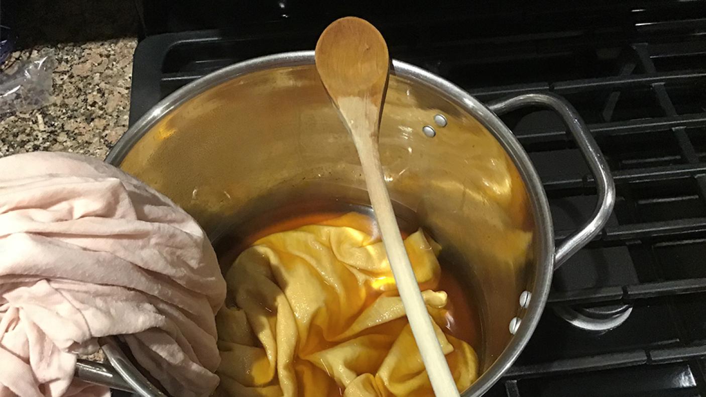 Pale pink shirt submerged halfway into amber dye in a stainless steel pot.
