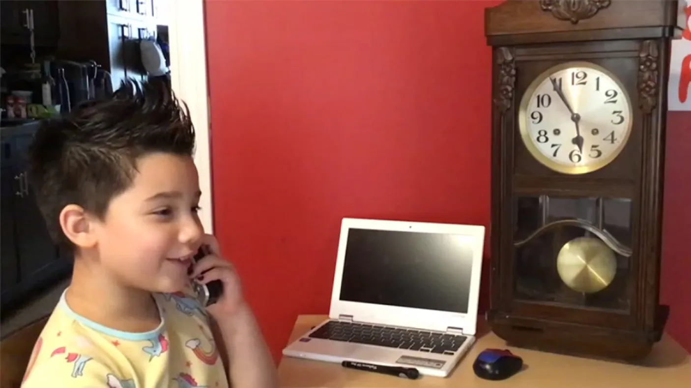 A girl with short spiky hair smiles while talking on the phone and sitting next to a laptop and grandfather clock on the table.
