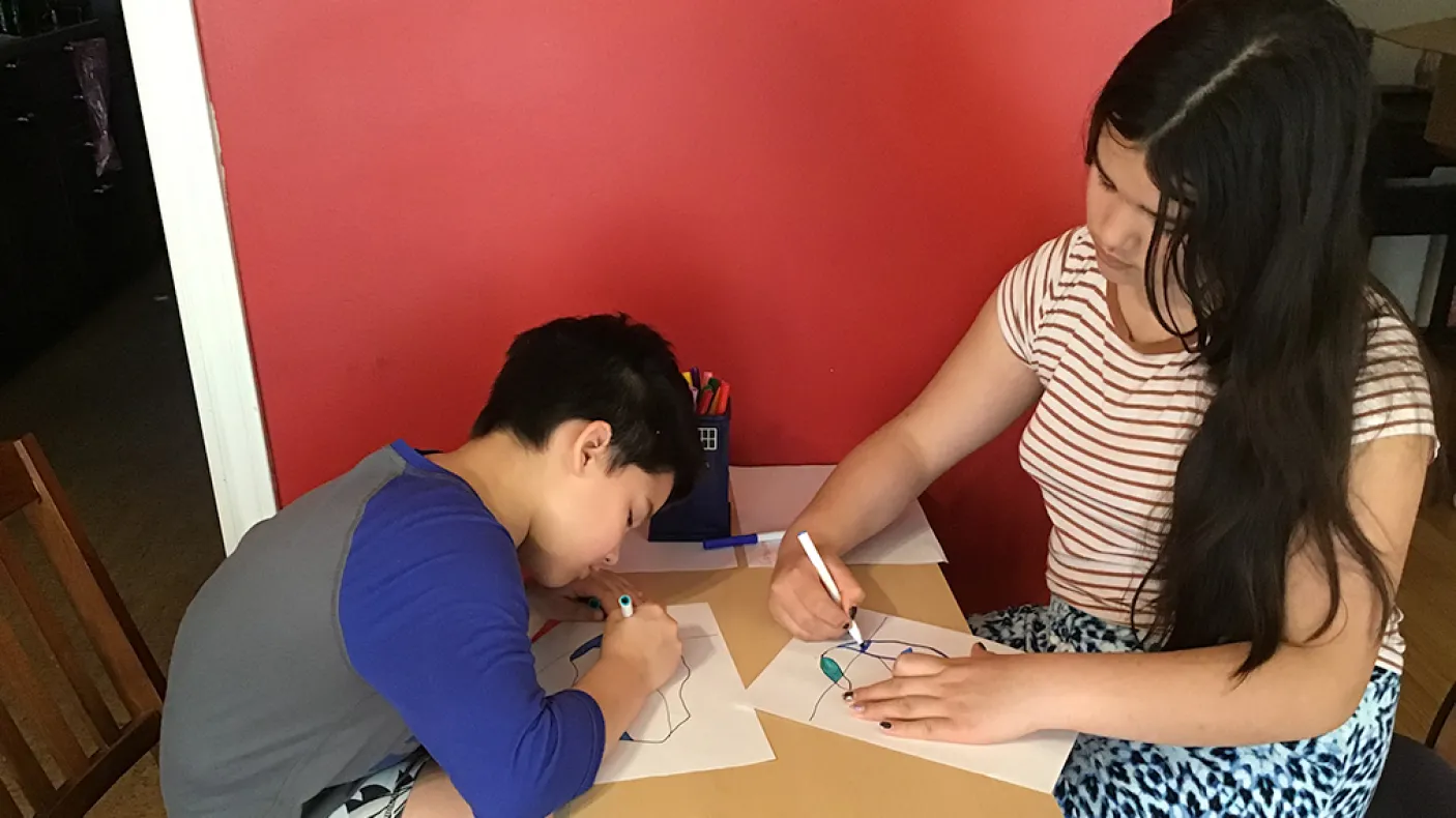 Deux filles, l'une aux cheveux courts et foncés et l'autre aux cheveux longs et foncés, s'assoient à une table et colorient des dessins d'icebergs avec des marqueurs.
