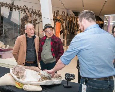 Deux hommes écoutent attentivement un expert en ROM leur parler d'un fossile de dinosaure.