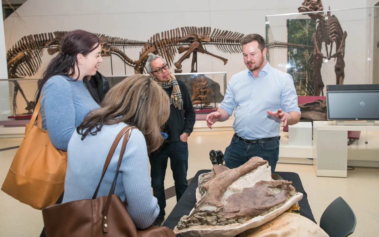 Trois personnes regardent un os de dinosaure pendant qu'un expert ROM explique.