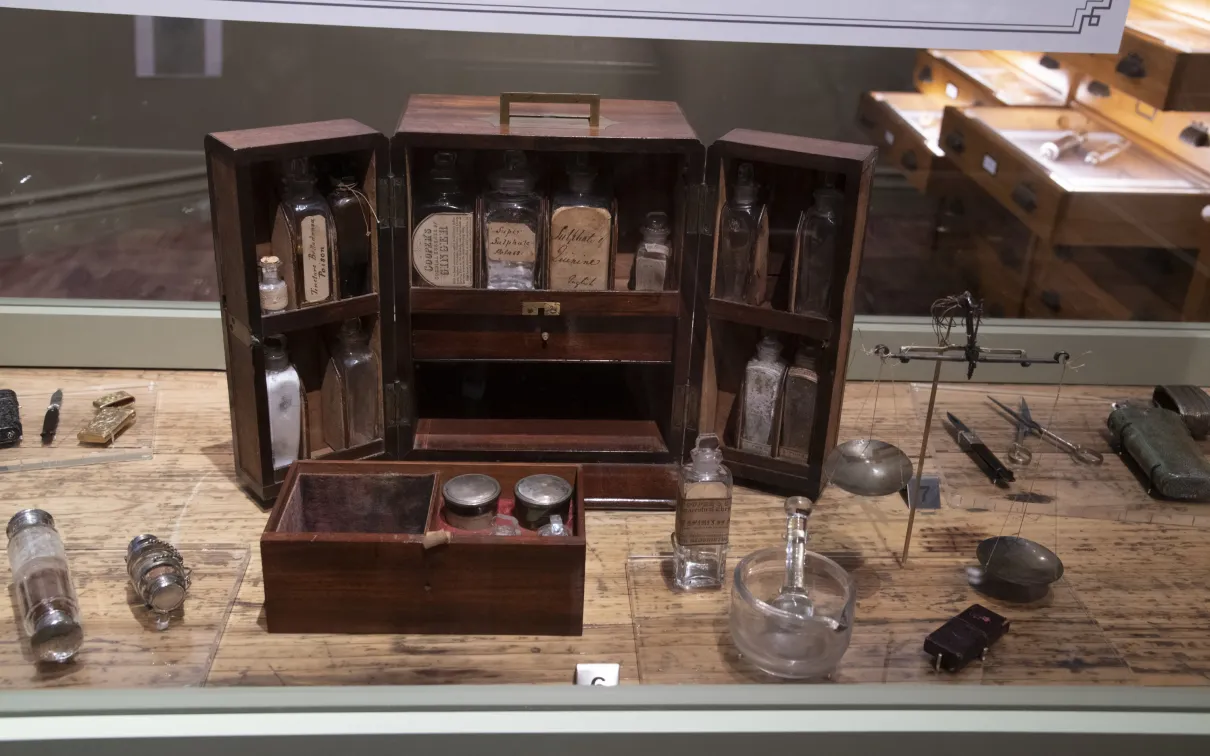 A vintage wooden apothecary box with glass bottles and containers is displayed on a wooden table alongside various old medical tools.