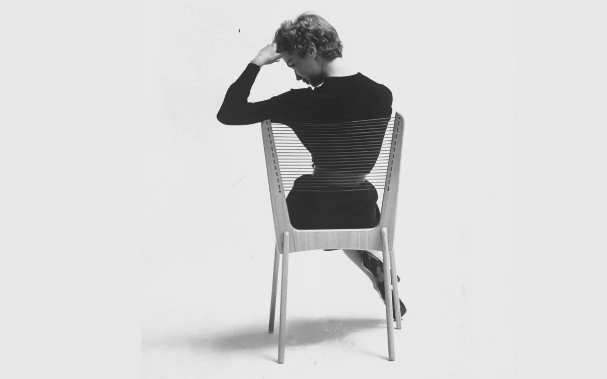 Monochrome 1950s image of the back of a woman in a black dress seated on a chair made of a wood frame and cord strung seat and back.