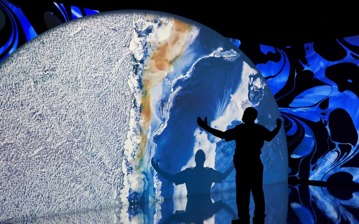 Projection of a planet with a silhouette of a man with his hands in the air looking at a large projection of a planet