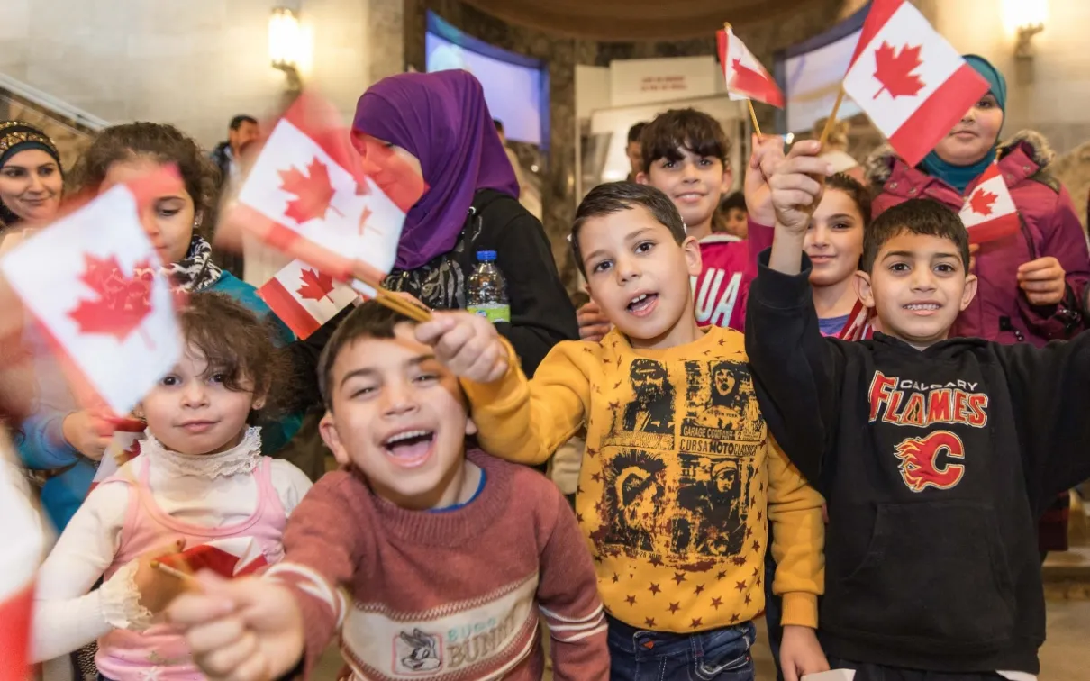 Un groupe d'enfants syriens sourit et agite des drapeaux canadiens devant la Galerie Schad de la biodiversité du ROM.
