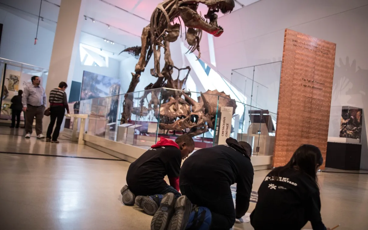 Children in the ROM in My Backyard program fill out an activity sheet in front of the T-rex at the ROM.