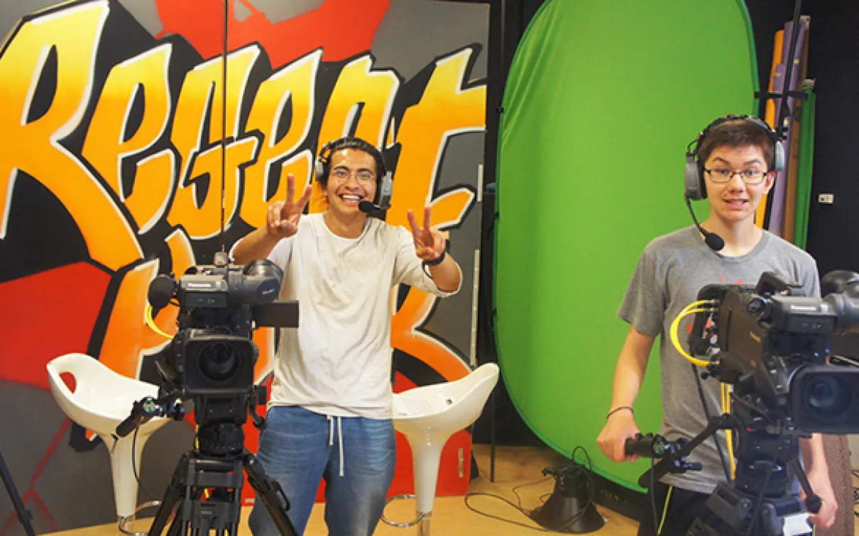 Two Youth Cabinet members wear headsets and stand behind video cameras.
