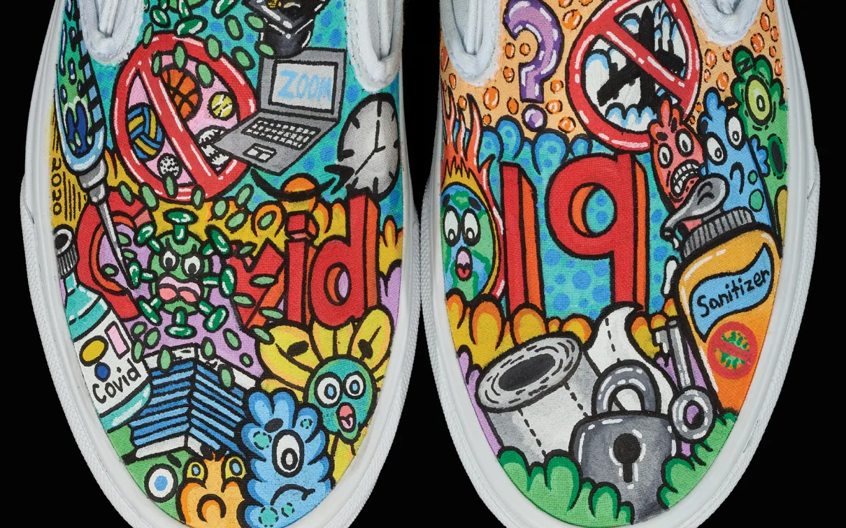 Image detail of a pair of white, slip-on sneakers with the tops painted in a colourful mosaic of COVID images and symbols.