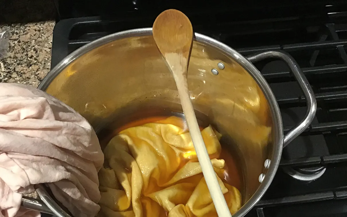 Chemise rose pâle immergée à moitié dans une teinture ambrée dans un pot en acier inoxydable.
