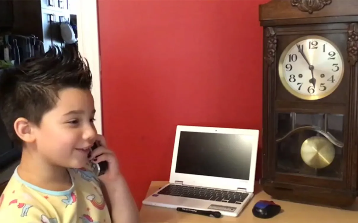 A girl with short spiky hair smiles while talking on the phone and sitting next to a laptop and grandfather clock on the table.
