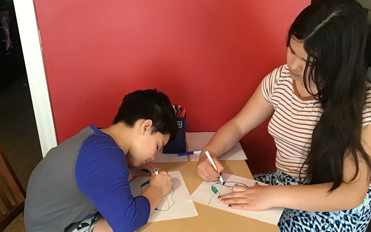 Deux filles, l'une aux cheveux courts et foncés et l'autre aux cheveux longs et foncés, s'assoient à une table et colorient des dessins d'icebergs avec des marqueurs.
