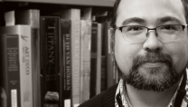 Gros plan en noir et blanc d'un homme portant une barbe et des lunettes devant une étagère de bibliothèque.