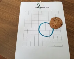 Biscuit aux pépites de chocolat sur papier quadrillé, cure-dent et trombone sur le papier, avec un cercle dessiné à partir du contour du biscuit à côté.