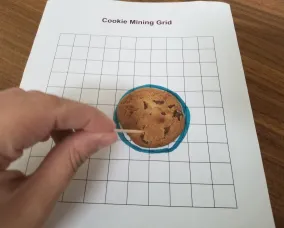 Chocolate chip cookie on grid paper in circle, toothpick poised to begin mining. Hand visible to begin the mining process